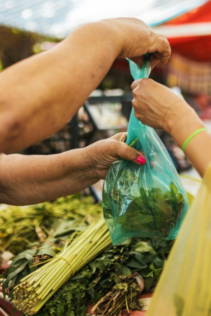 A close-up of a fresh produce exchange at an outdoor market with hands exchanging a bag of greens.